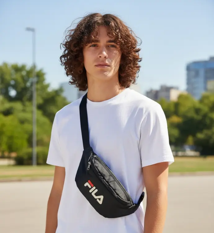 Black Fila crossbody bag (or fanny pack) with the red, white, and black Fila logo, worn across the chest by a person in a white t-shirt, perfect for casual or street style.